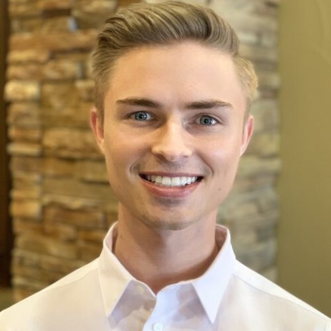 Young man in white dress shirt smiling at camera in dental office setting.